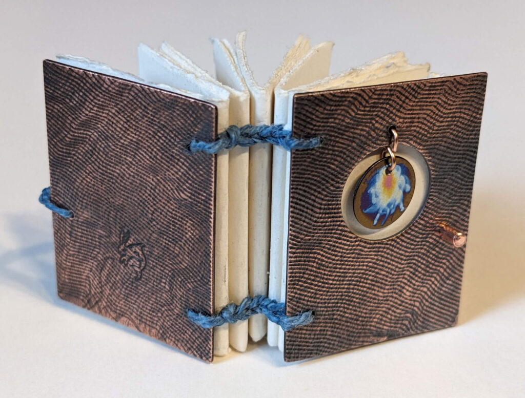 Small handmade book with textured copper covers and a circular piece of niobium with an abstract design in rainbow colors.