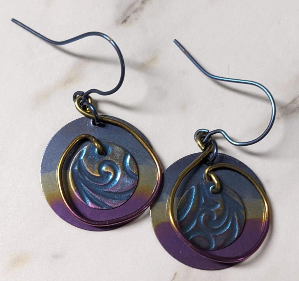 Two colorful earrings with swirls of wire holding textured metal disks over larger disks with stripes of color.